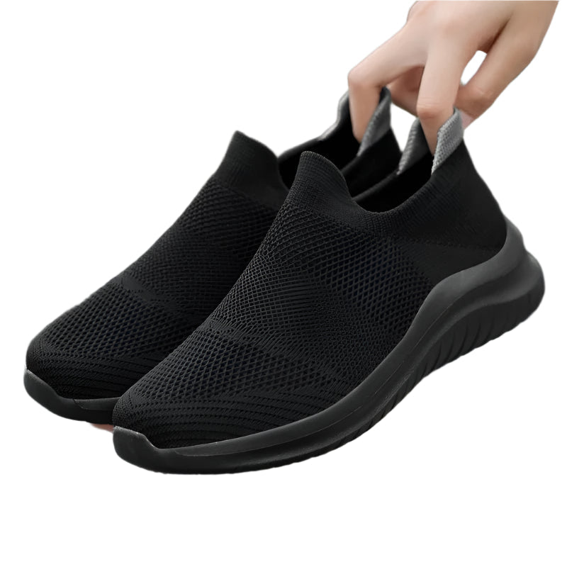 Black slip-on sneakers being held by a hand on a white background