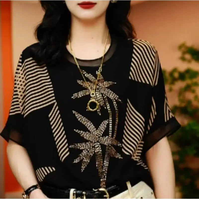 Woman wearing a black top with gold patterns and a necklace, against a blurred background.