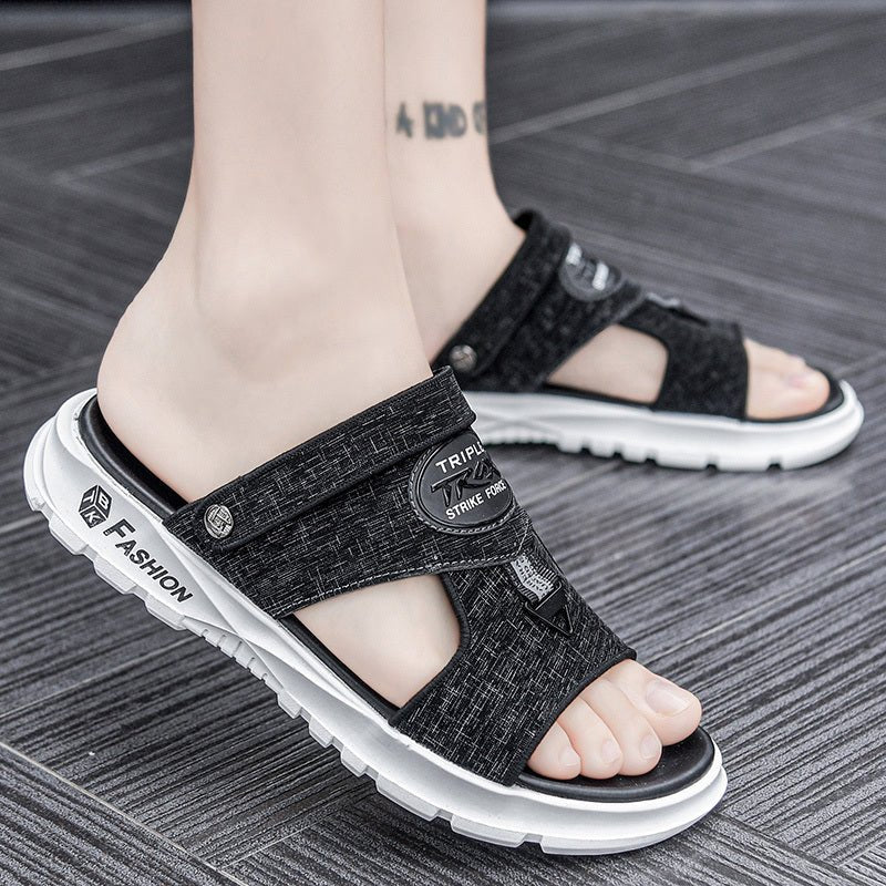 Black sandals with white soles worn by a person on a wooden floor.