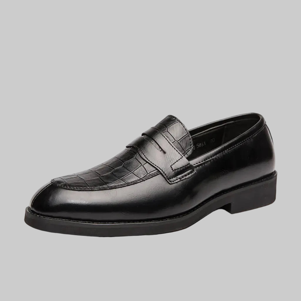 A man's foot wearing a brown crocodile-patterned leather shoe with a buckle strap, alongside a wooden floor and a wristwatch.