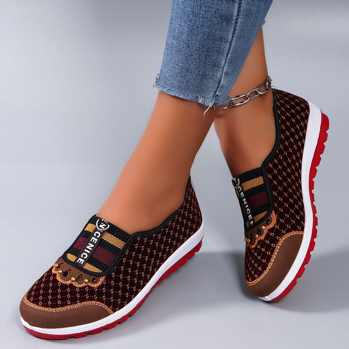 Brown patterned slip-on shoes with red soles worn by a person in jeans on a gray background