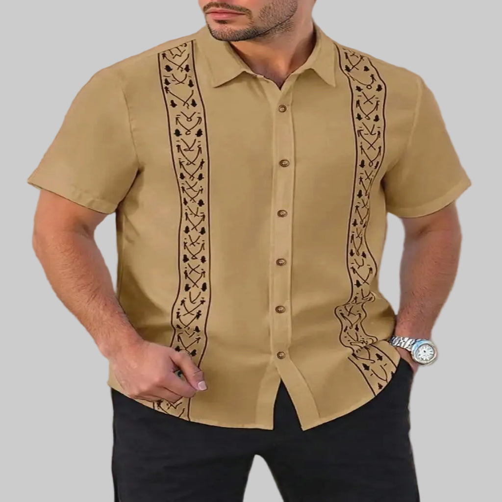 Man wearing a beige short-sleeve guayabera shirt with black embroidery against a beige wall.
