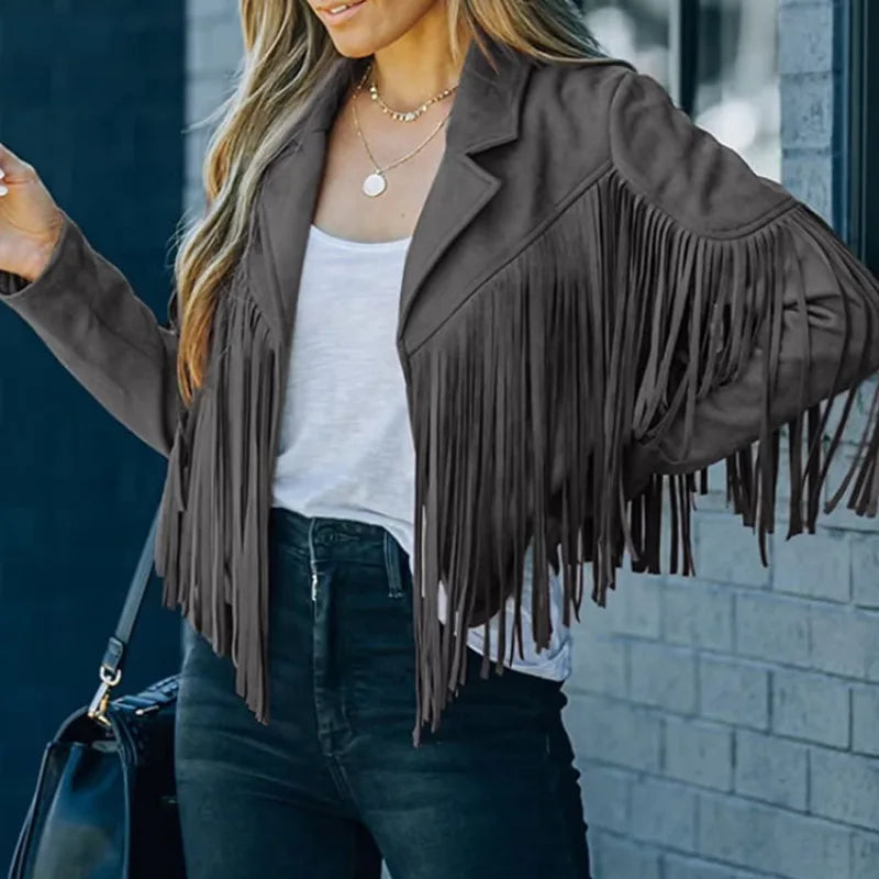 Person wearing a brown fringe jacket with a white top and dark jeans, standing against a brick wall.