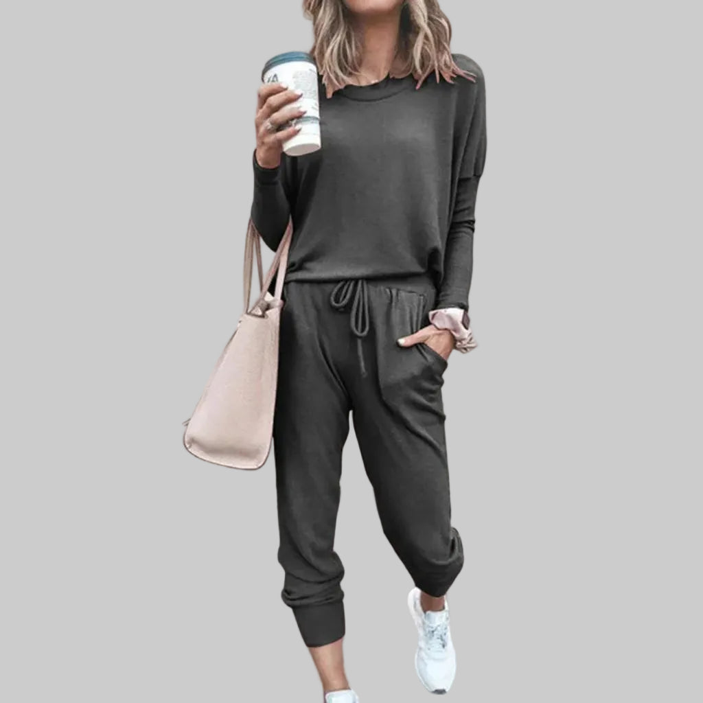 Person wearing a dark gray tracksuit with a beige handbag and white sneakers, standing against a light pink wall.