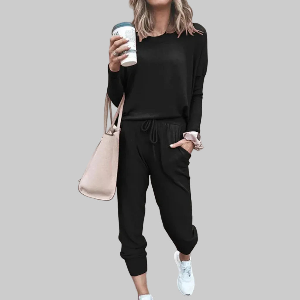 Person wearing a black outfit with a beige handbag and white sneakers, standing against a light pink wall.
