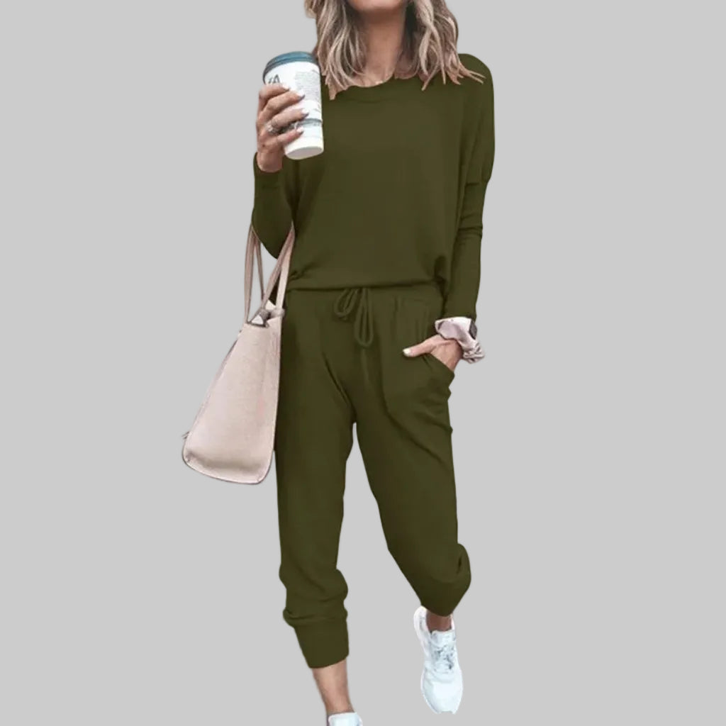 Person wearing an olive green outfit with a beige bag and white sneakers, standing against a light gray background.
