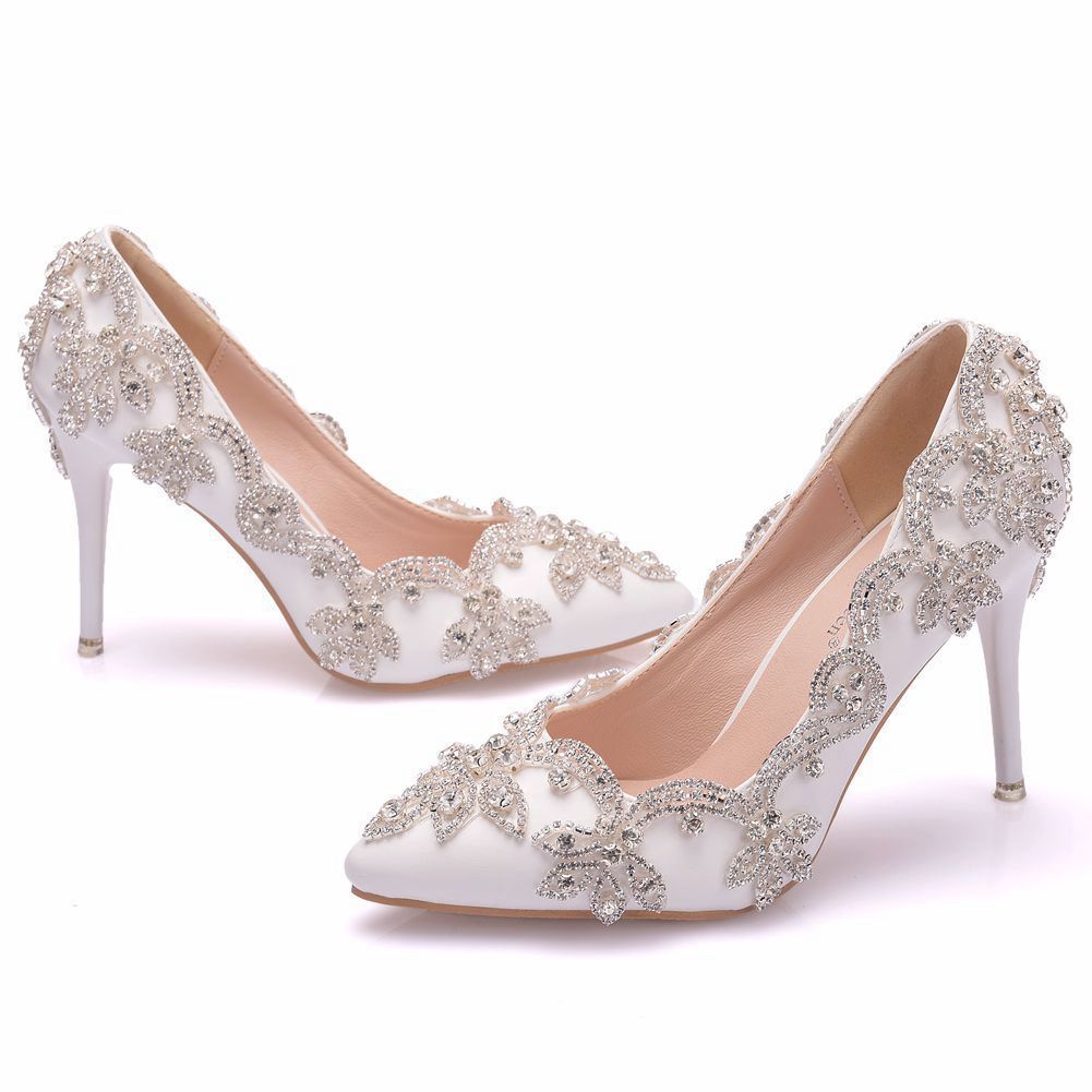 A pair of white high heel shoes with rhinestone details, displayed in a home setting. The shoes are pointed-toe and have a stiletto heel design.